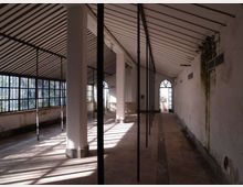 Interno di un lungo spazio industriale dismesso con soffitto a falde e travi a vista, pilastri bianchi e sottili montanti metallici in fila. Lungo il lato sinistro ci sono ampie finestre a griglia che lasciano entrare luce radente, mentre sul fondo si vede una porta ad arco.