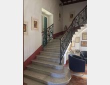 Interno di un edificio con ampia scalinata in marmo che curva verso l’alto, con ringhiere in ferro battuto decorato. Pareti bianche con fascia rossa, quadri e cornici alle pareti, una finestra con persiane verdi e un piccolo angolo salotto con poltrone e tavolino.