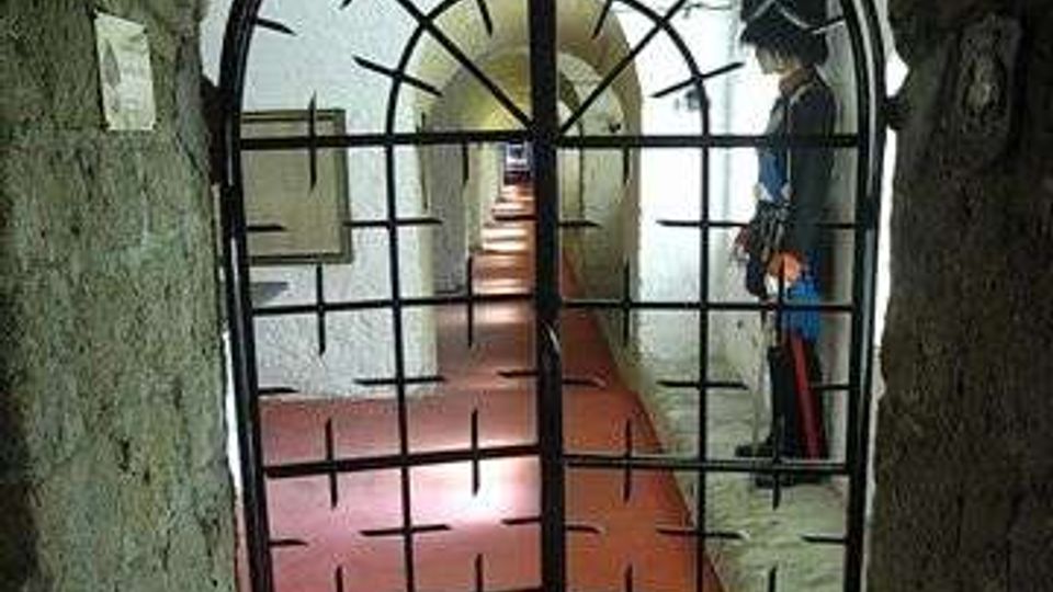 Corridoio sotterraneo con volta in mattoni e pavimento rossastro, visto attraverso un cancello in ferro ad arco. In fondo si intravedono pareti bianche illuminate e una serie di passaggi ad arco lungo il tunnel.