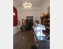 Interno di un bar stretto e allungato con pavimento in marmo, bancone in acciaio con vetrina e scaffali in legno con bicchieri e bottiglie. Lampadario classico a soffitto, pareti bianche con una sezione rosso scuro e porta a vetri sul fondo.