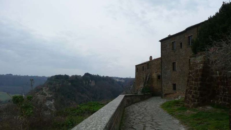 Sentiero lastricato in pietra con muretto di parapetto che costeggia un edificio in muratura su un crinale. Sullo sfondo si vedono colline e una valle boscosa sotto un cielo nuvoloso.