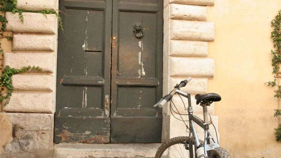 Ingresso di un edificio con grande portone in legno scuro sotto un arco in pietra bugnata e grata semicircolare in ferro, con edera che ricopre parte della facciata. Davanti al portone, su pavimentazione in sampietrini, è appoggiata una bicicletta.