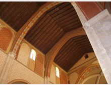 Interno di una chiesa con soffitto a capriate in legno scuro e grandi archi in mattoni decorati con motivi geometrici. Pareti in pietra chiara con finestre ad arco che lasciano entrare luce naturale.