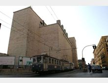 Grande edificio urbano in mattoni chiari con facciate quasi cieche e volumi squadrati, affacciato su una strada con binari del tram. In primo piano passa un tram sotto una rete di cavi elettrici; sul lato destro si vedono altri palazzi e un incrocio con semaforo.