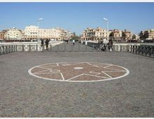 Ampio ponte pedonale pavimentato in pietra con una grande stella circolare decorativa al centro, delimitato da ringhiere metalliche e lampioni. Sullo sfondo si vedono edifici urbani lungo il waterfront sotto un cielo sereno.