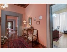 Corridoio interno di una casa con pareti color salmone, pavimento in parquet e tappeto persiano; lungo la parete c’è una consolle decorata con specchio, lampade e quadretti. Porte con cornici grigie si aprono su altre stanze, tra cui un soggiorno luminoso con grandi tende e una lampada a sospensione.