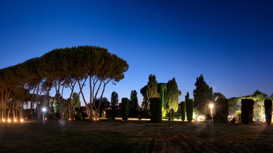 Ampio parco al crepuscolo con prato in primo piano e una fila di alti pini a ombrello sulla sinistra, affiancati da cipressi e siepi potate. Luci a terra e lampioni illuminano i vialetti e alcune strutture sullo sfondo sotto un cielo blu scuro.