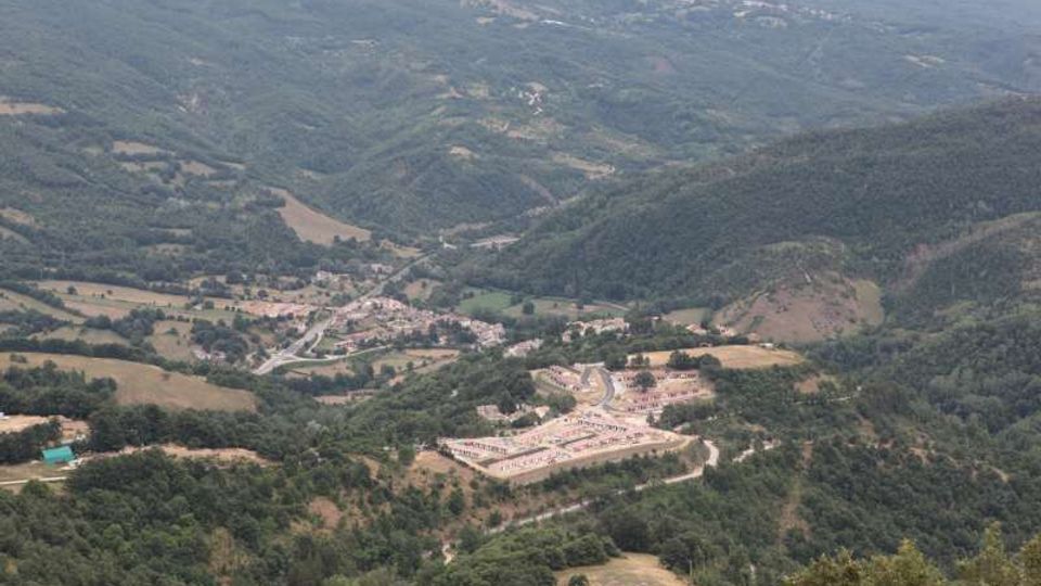 Veduta panoramica di una valle montuosa con colline boscose e campi coltivati, attraversata da strade che collegano un piccolo paese. In primo piano si vede un ampio complesso con piazzale e parcheggi, circondato dal verde.
