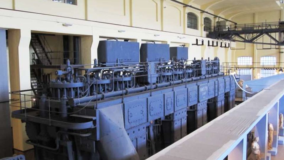 Grande sala industriale con soffitto alto ad arcate e finestre ad arco, vista da una passerella sopraelevata con ringhiere. Al centro si trova un enorme macchinario blu con tubazioni e pannelli metallici, tipico di una centrale o impianto storico.