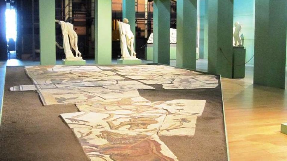Interno di un museo in un ambiente industriale con passerelle metalliche e tubazioni a vista, dove una lunga pavimentazione a mosaico frammentata è esposta al centro. Strutture verdi con aperture incorniciano l’area espositiva e ospitano statue su piedistalli lungo il percorso.