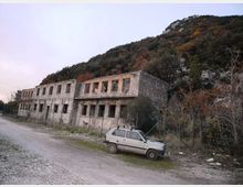 Edificio rettangolare in muratura grezza a due piani, con file di finestre vuote e portico frontale, situato lungo una strada sterrata. Sullo sfondo si vede un pendio collinare boscoso e roccioso, con vegetazione fitta e recinzione metallica vicino alla struttura.