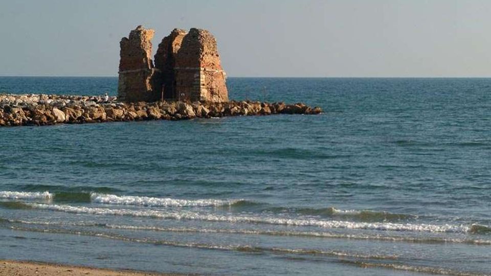 Resti di una torre in muratura su un piccolo molo di rocce che si protende nel mare, con spiaggia sabbiosa e onde basse in primo piano. Sullo sfondo si vede l’orizzonte marino sotto un cielo sereno.