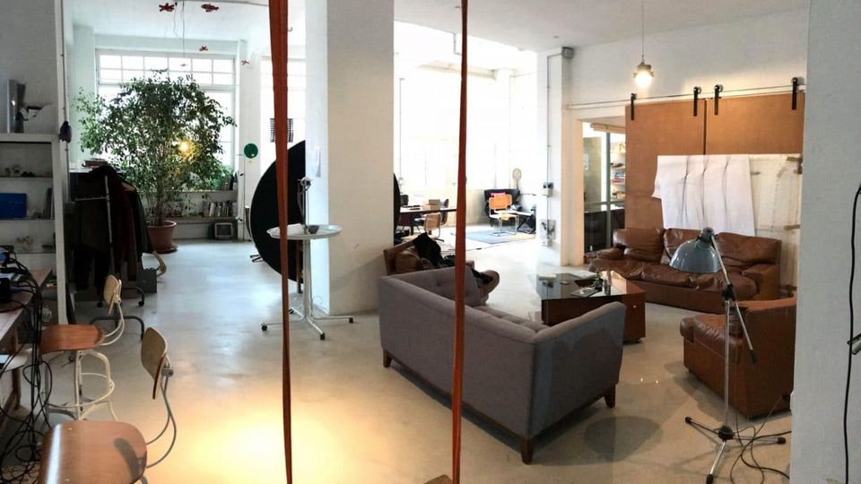 Ampio loft open space con grandi finestre e pilastri bianchi, con area salotto composta da divani in pelle e in tessuto, tavolino e lampade. Sullo sfondo si vedono postazioni di lavoro, scaffali e una grande pianta in vaso, con una porta scorrevole su binario.
