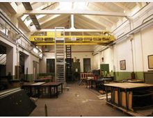 Interno di un capannone industriale adibito a officina, con soffitto alto a capriate e lucernari che illuminano lo spazio. Al centro è presente un carroponte giallo e una scala metallica, con banchi da lavoro e macchinari lungo le pareti.