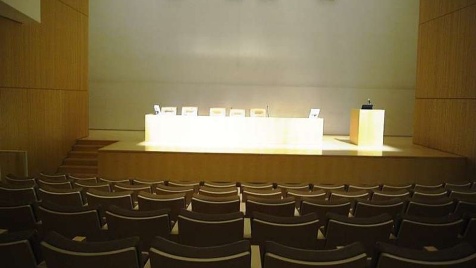 Sala conferenze moderna con file di poltroncine rivolte verso un palco rialzato, con lungo tavolo bianco per relatori e podio laterale. Pareti rivestite in legno e parete di fondo chiara con piccole aperture quadrate in alto.