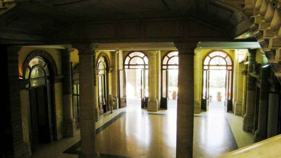 Atrio interno di un edificio storico con colonne in pietra, pavimento chiaro a riquadri e più porte ad arco vetrate che danno verso l’esterno. In alto si vedono balaustre e un’ampia scala in pietra lungo il lato destro.