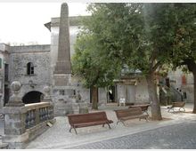 Piccola piazza lastricata con panchine in legno e alberi ombrosi, dominata da un alto obelisco monumentale su basamento in pietra accanto a una balaustra. Sullo sfondo si vedono edifici in muratura e intonaco con arcate e ingressi al piano terra.