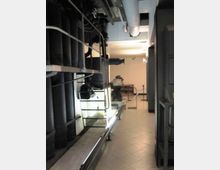 Interno di una sala tecnica o museo industriale con grandi macchinari metallici e tubazioni lungo le pareti e il soffitto, illuminata da luci lineari. Il pavimento è piastrellato e un corridoio centrale conduce verso altre apparecchiature esposte in fondo.