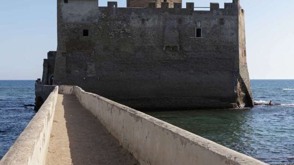 Fortezza in pietra sul mare con mura merlate e torre centrale, raggiungibile tramite una stretta passerella con parapetti in cemento. Il castello sorge su uno scoglio circondato dall’acqua, con cielo sereno sullo sfondo.