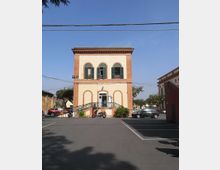 Edificio pubblico a due piani in stile tradizionale, con facciata color crema e cornici in mattoni, finestre ad arco con persiane verdi e ingresso centrale raggiungibile da una rampa con ringhiera. Davanti si apre un piazzale asfaltato con parcheggi e cavi aerei, circondato da alcuni alberi e costruzioni laterali.