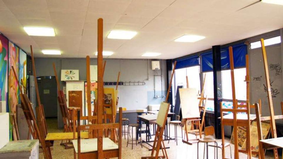 Aula d’arte con numerosi cavalletti e tele, tavoli e sgabelli disposti nello spazio su pavimento in graniglia. Grandi finestre laterali con tende blu e pannelli murali colorati lungo una parete.