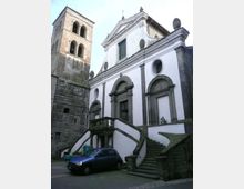 Facciata di una chiesa in stile barocco con intonaco bianco e cornici in pietra scura, ingresso sopraelevato raggiunto da una scalinata doppia curva con balaustra. A sinistra si vede un campanile in pietra con finestre ad arco, in una piccola piazza pavimentata.