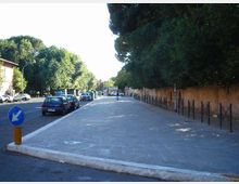 Ampio marciapiede pavimentato lungo una strada urbana con auto parcheggiate a sinistra e una lunga fila di alberi e recinzione metallica sul lato destro. Sullo sfondo si vedono edifici bassi e segnaletica stradale, in un contesto di viale alberato.