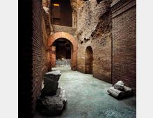 Interno di un sito archeologico con corridoio in mattoni e pietra, pareti alte con nicchie e un grande arco in laterizio che conduce a un’area più ampia. Sul pavimento in cemento si vedono blocchi di pietra e frammenti di rovine.