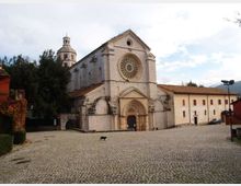 Facciata di una grande chiesa in pietra chiara su una piazza lastricata, con portale ad arco e grande rosone circolare centrale. Sullo sfondo si vedono un campanile e edifici annessi con tetti in coppi, circondati da alberi.