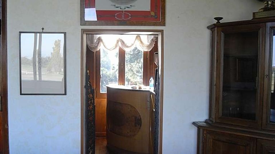 Interno di una sala con tavolo e sedie in legno intagliato, una credenza vetrata sulla destra e quadri appesi a una parete chiara. Al centro, una porta aperta conduce a un’area illuminata con finestra e tende, con vista sul verde esterno.