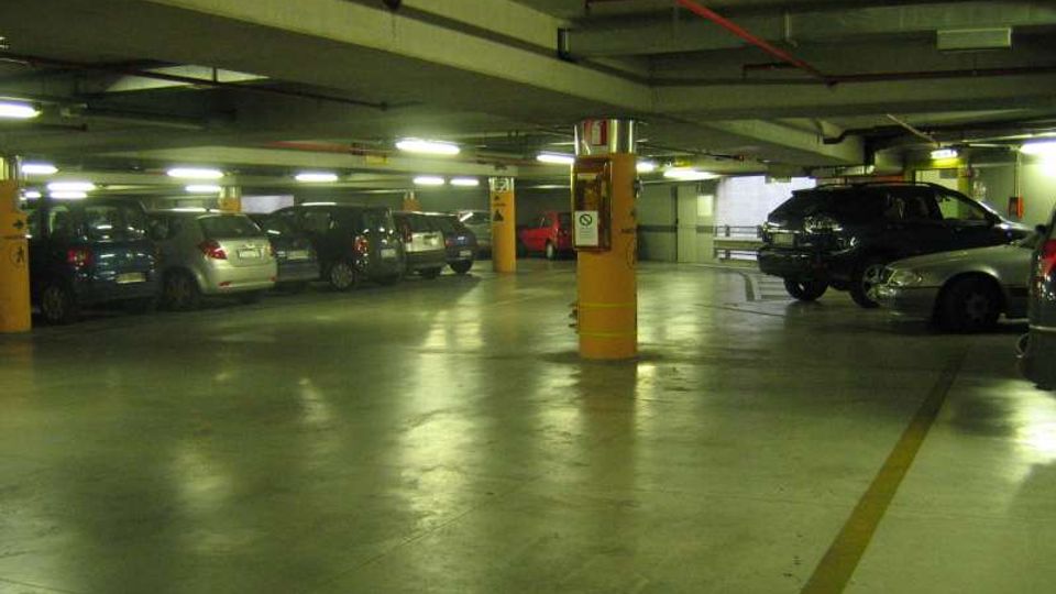 Parcheggio sotterraneo con pavimento in cemento lucido, colonne arancioni e illuminazione al neon sul soffitto con tubazioni a vista. Auto parcheggiate lungo i lati e corsia centrale libera delimitata da linee gialle.