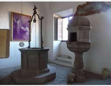 Interno di una piccola cappella o museo con pareti bianche e pavimento in pietra, con un pozzo in pietra dotato di struttura in ferro al centro. A destra c’è un fonte battesimale scolpito con cupola decorata e una finestra aperta che illumina l’ambiente; sulla parete si vede un grande dipinto religioso incorniciato.