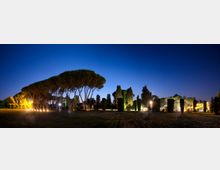 Ampio parco al crepuscolo con prato in primo piano e una fila di alti pini a ombrello sulla sinistra, affiancati da cipressi e siepi potate. Luci a terra e lampioni illuminano i vialetti e alcune strutture sullo sfondo sotto un cielo blu scuro.