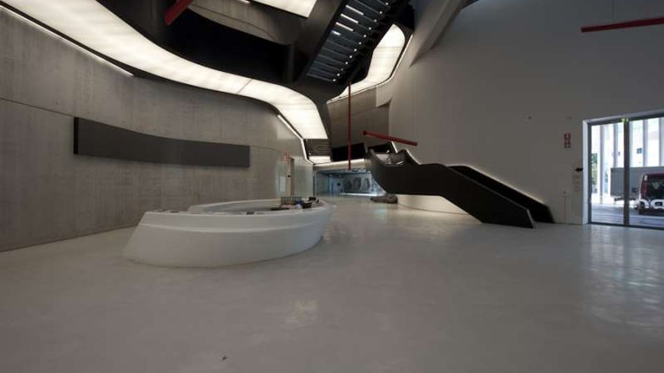 Ampio atrio interno di un edificio moderno, con pavimento chiaro, pareti in cemento e un banco reception bianco curvo al centro. Una grande scala nera a rampe e passerelle sospese attraversa lo spazio sotto un soffitto con pannelli luminosi curvilinei, con ingresso vetrato sul lato destro.