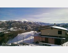 Casa di montagna con tetto innevato e recinzione metallica in primo piano, su un pendio coperto di neve. Sullo sfondo si vede un’ampia catena di montagne con cime innevate sotto un cielo sereno.