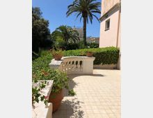 Terrazza piastrellata con balaustra in pietra chiara e vasi di fiori, affacciata su un giardino con siepi e alte palme. Sullo sfondo si vedono edifici chiari e cielo sereno.