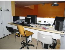 Piccola sala di controllo con banco attrezzato con mixer audio, computer e monitor, cavi e altoparlanti. Sono presenti sedie e armadi laterali, con pareti interne in legno e arancione e un proiettore fissato al soffitto.