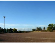 Campo sportivo all’aperto con superficie in terra battuta, una porta da calcio e pali per l’illuminazione. Sullo sfondo si vedono recinzioni alte e alberi sotto un cielo sereno.