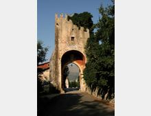 Antica porta d’accesso in pietra con torre merlata e grande arco che sovrasta una strada, con una piccola finestra e segni di intonaco consumato. Alberi e vegetazione fiancheggiano l’arco e la carreggiata, con colline visibili sullo sfondo.