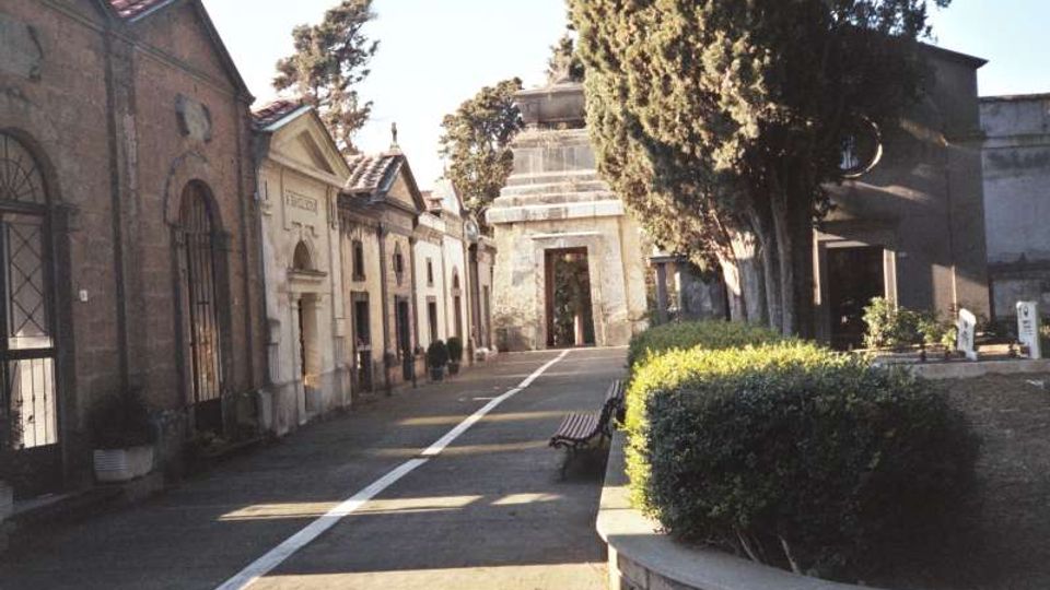 Viale pavimentato in un cimitero, affiancato da cappelle funerarie in muratura con facciate ad arco e portali. A destra ci sono siepi potate e alti cipressi, con un mausoleo in pietra sullo sfondo.