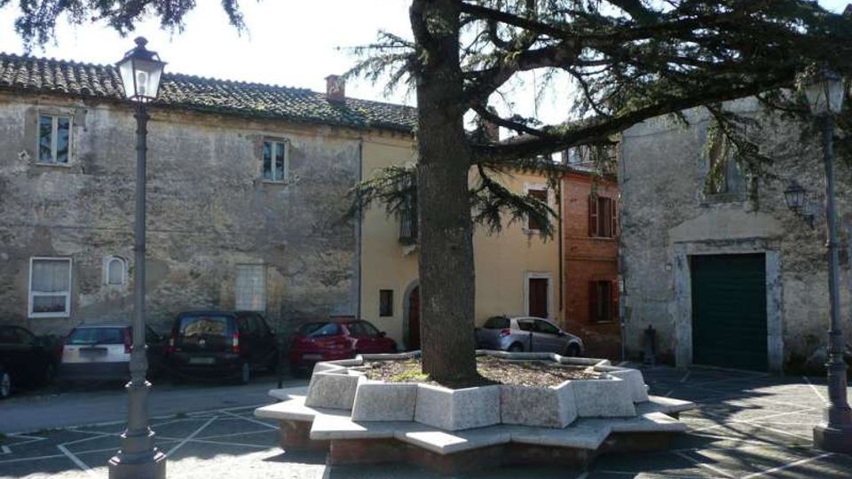 Piccola piazza lastricata con al centro un grande albero sempreverde circondato da una panchina e aiuola in pietra poligonale. Sullo sfondo ci sono edifici in muratura intonacata con finestre e portoni, alcune auto parcheggiate e lampioni in stile tradizionale.