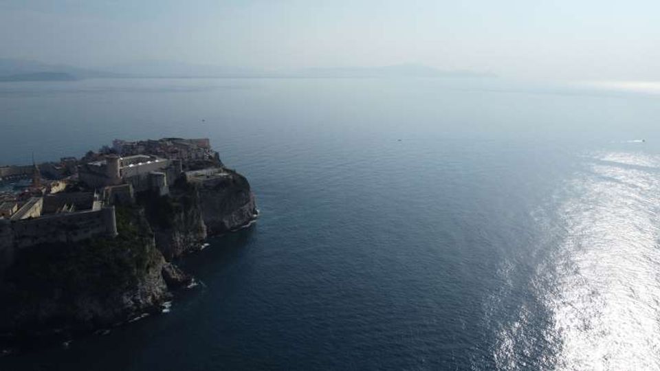 Veduta aerea di una città fortificata su un promontorio roccioso a picco sul mare, con mura e edifici compatti lungo la scogliera. Ampia distesa d’acqua calma con riflessi del sole e costa lontana appena visibile all’orizzonte.