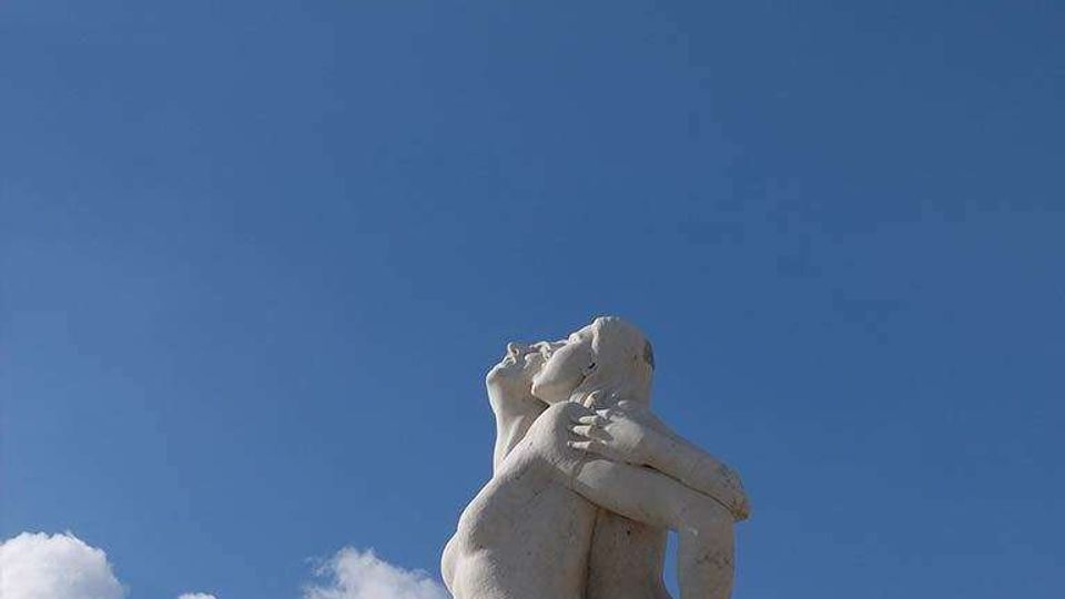 Statua in marmo di due figure nude abbracciate su un piedistallo, ripresa dal basso contro un cielo azzurro con alcune nuvole. Ambiente esterno con ampio sfondo di cielo.
