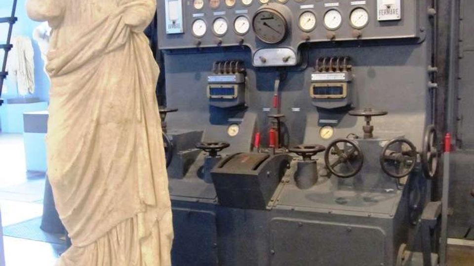 Interno industriale con grande pannello di controllo in metallo, pieno di manometri, leve e volantini, collegato a tubazioni scure. In primo piano, una statua classica senza testa su piedistallo bianco, in uno spazio tipo museo o impianto storico.