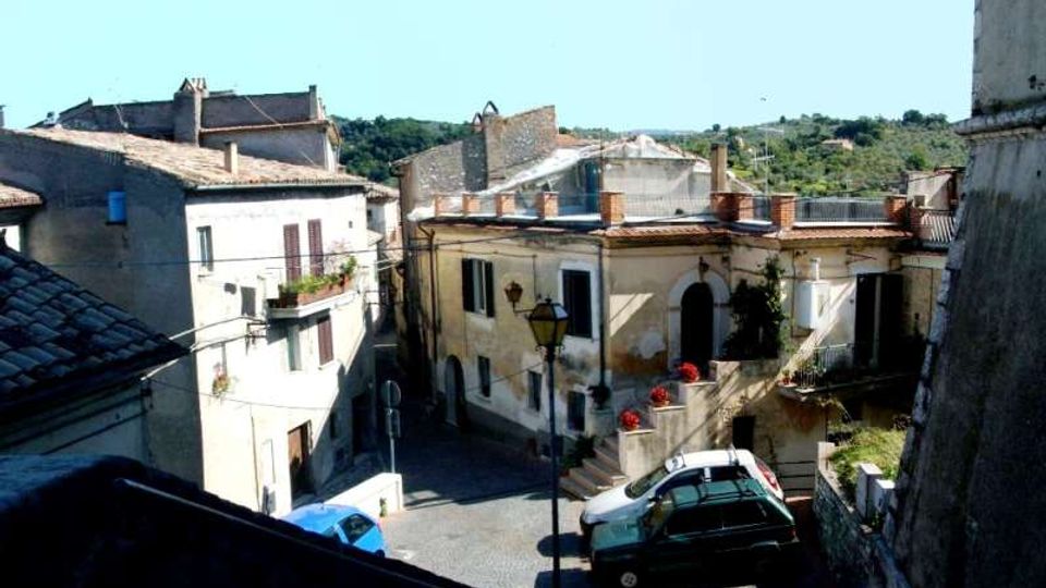 Veduta dall’alto di una piccola piazza in un borgo con case in muratura intonacata e tetti in tegole, balconi e finestre con persiane. Una strada in pendenza con lampione e auto parcheggiate attraversa lo spazio, con colline verdi sullo sfondo.