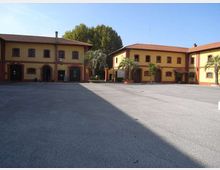 Cortile pavimentato e ampio circondato da edifici a due piani in stile rurale, con facciate gialle e bordi rossi, tetti in tegole e arcate al piano terra. Sullo sfondo si vedono alberi alti e alcune palme vicino agli ingressi.