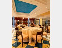 Sala ristorante elegante con tavoli rotondi apparecchiati con tovaglie chiare e sedie scure, su pavimento a motivi geometrici blu e gialli. Soffitto a livelli con riquadro centrale azzurro e faretti, colonne blu, pareti vetrate laterali e area bar sul fondo.