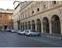 Strada urbana lastricata con auto parcheggiate lungo il marciapiede, affiancata da un grande edificio storico in pietra con porticato ad archi e cancellate in ferro. Sullo sfondo si vedono altri palazzi in stile classico con facciate ocra e finestre ornate.