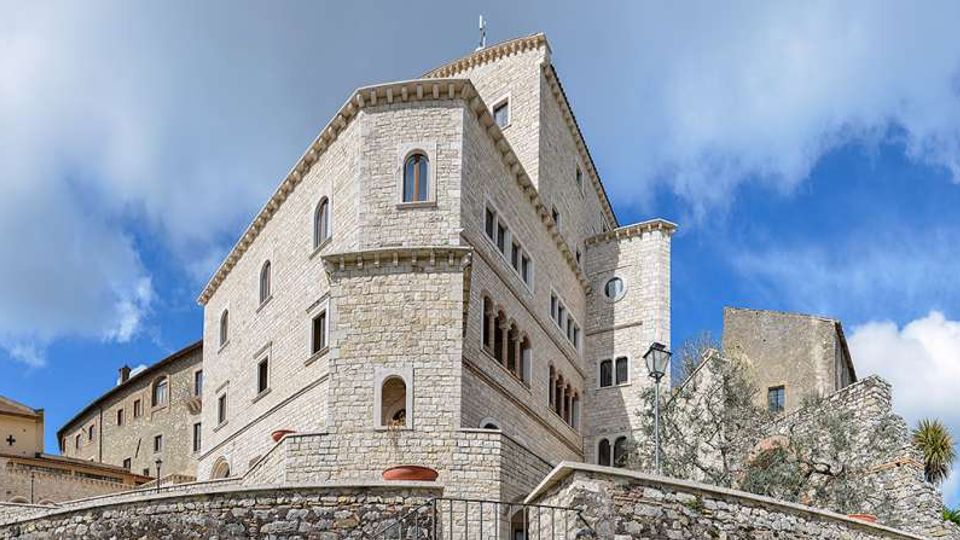 Grande edificio in pietra chiara in stile medievale, con finestre ad arco e una torre laterale, visto dal basso oltre un muro di cinta in pietra. Intorno si notano altri edifici storici e un lampione, sotto un cielo azzurro con nuvole.