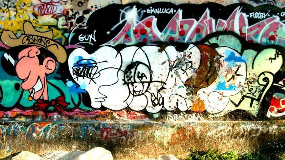 Muro di cemento all’aperto ricoperto da graffiti e scritte colorate, con grandi forme a lettere e un disegno in stile fumetto. Alla base del muro si vedono un marciapiede in cemento, ghiaia e alcune piante basse.